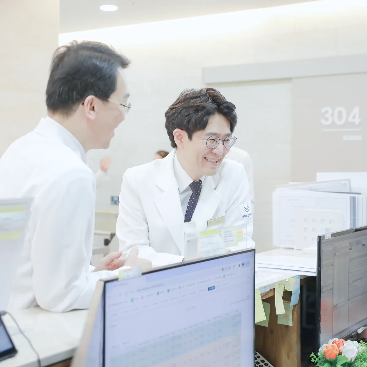 간호사실 모니터 앞에서 두 원장이 수술 후 회복 중인 환자의 기록을 보며 관리 계획을 상의하는 모습