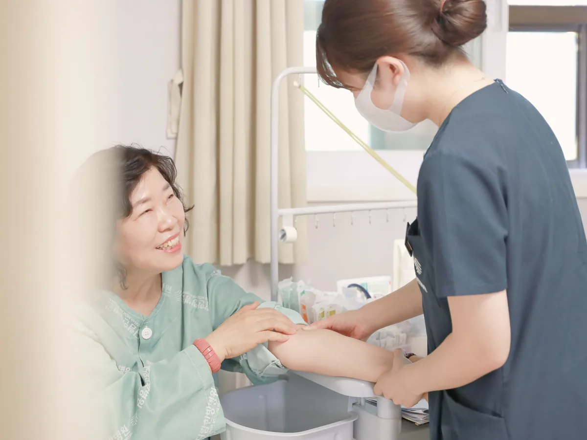 간호사가 환자 팔을 살피며 처치하는 동안 환자가 웃으며 이야기하는 병실 장면