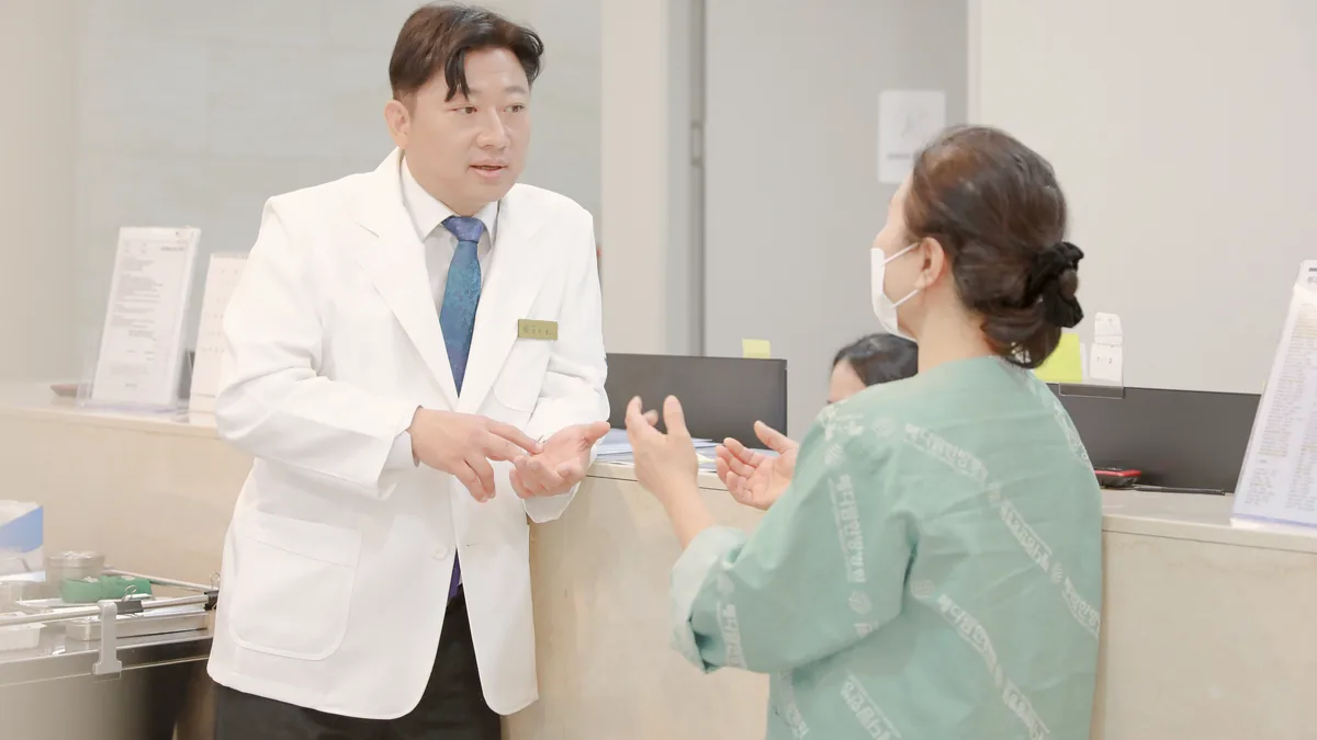 환자가 간호사실 앞에서 원장에게 손발 붓기와 숨찬 증상을 설명하며 상담하는 모습