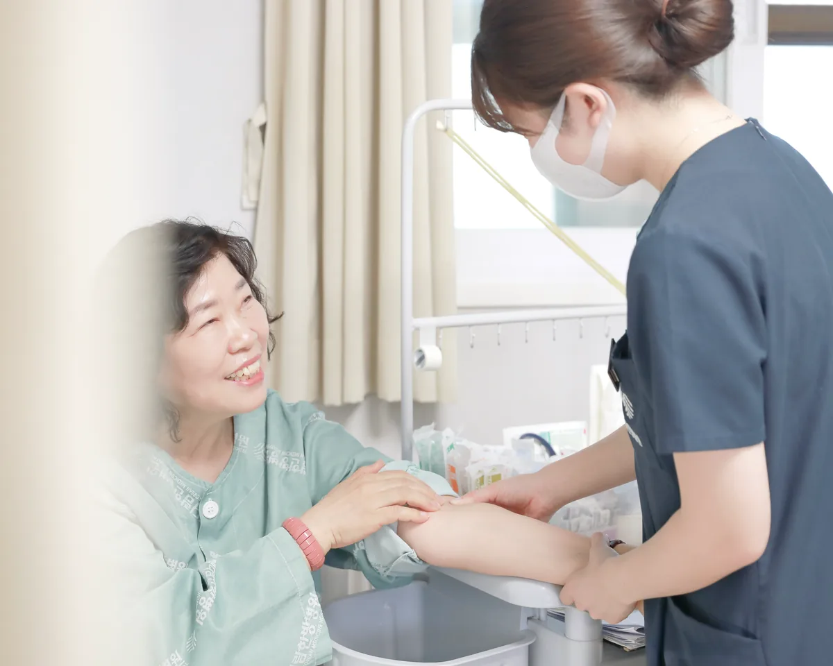 간호사가 환자 팔을 잡고 혈액 검사를 준비하는 동안 환자가 미소 짓고 있는 병실 장면