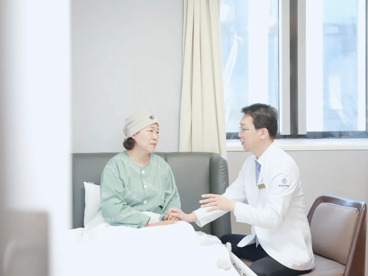 병실 침대 곁에 앉은 원장이 항암 치료 중인 환자의 손을 잡고 최근 생긴 증상을 차분히 묻는 모습
