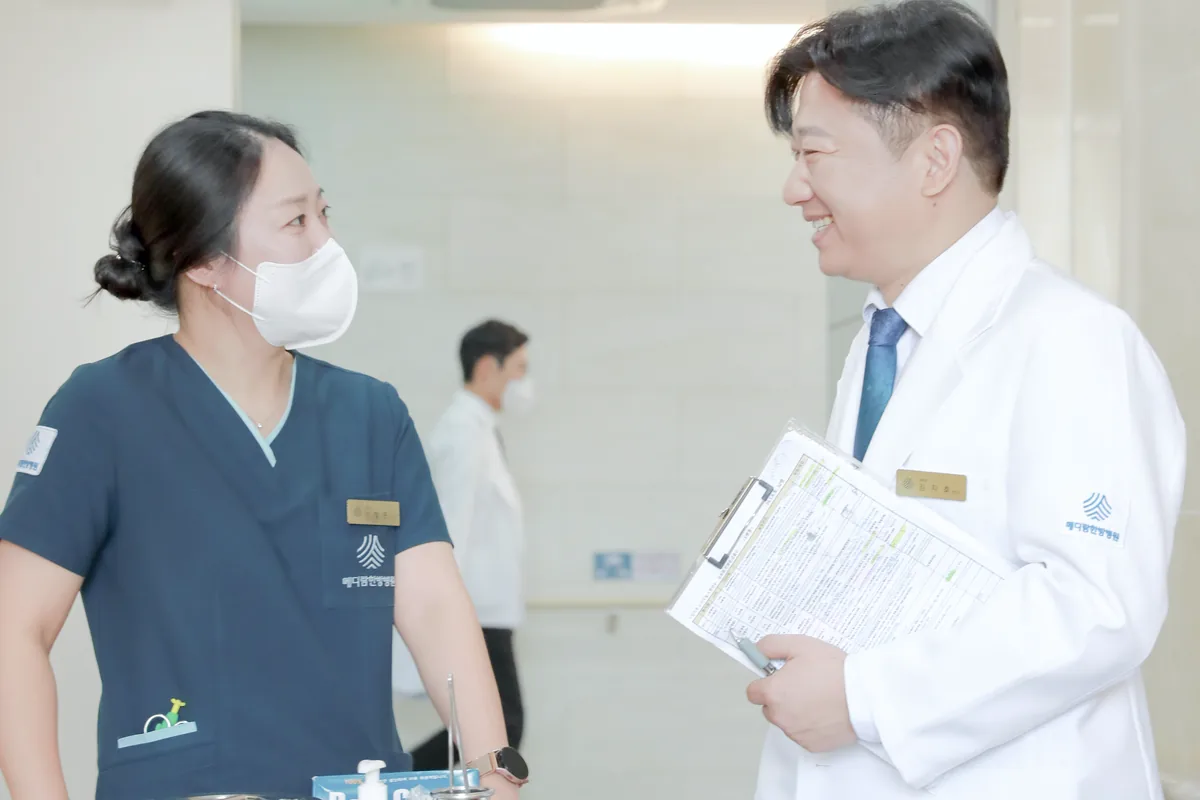 복도에서 원장이 간호사와 처치 카트 옆에 서서 항암 주사를 맞은 환자의 상태를 이야기하는 모습